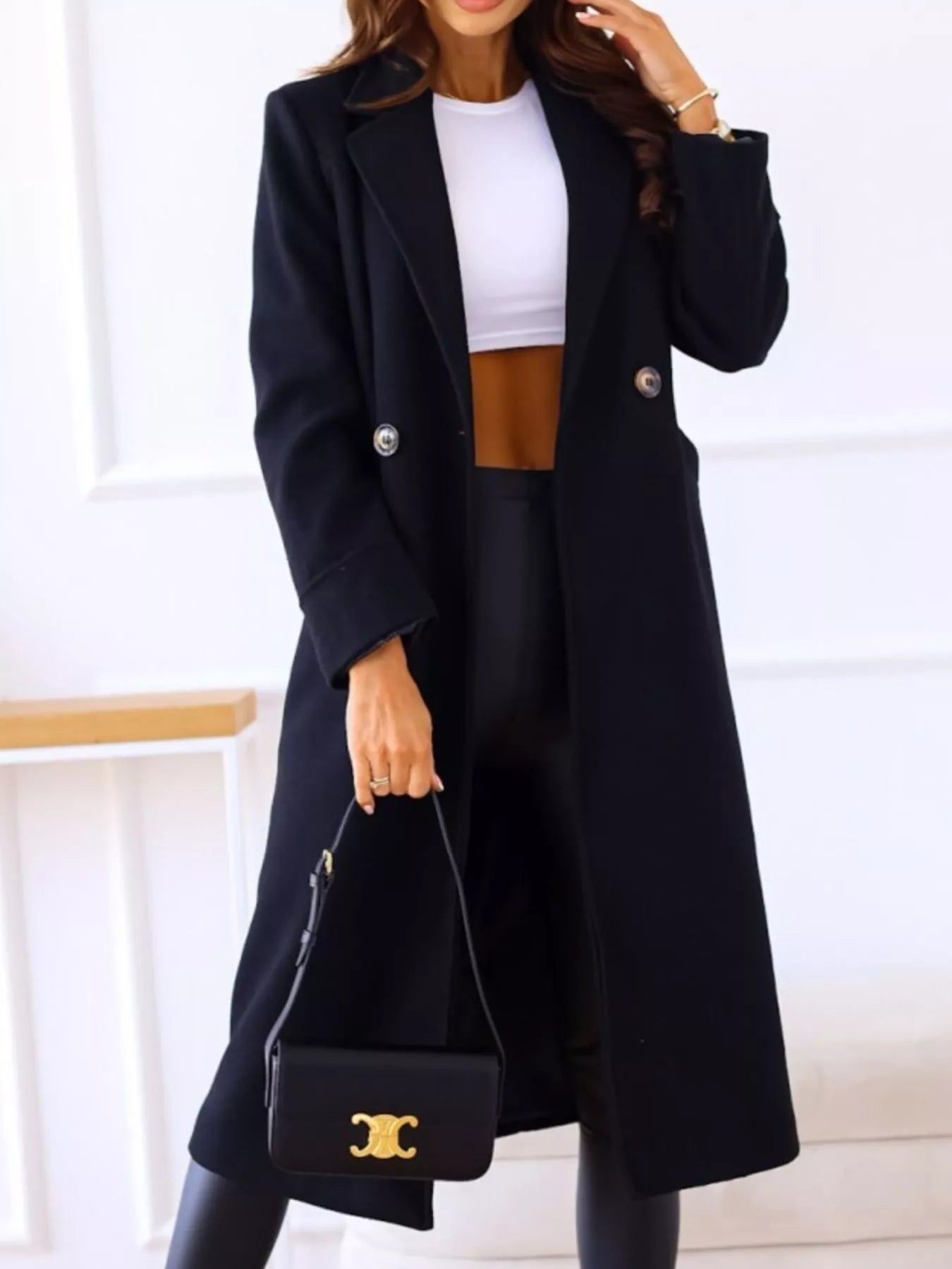Person wearing a black coat holding a black handbag with a gold logo on a white background