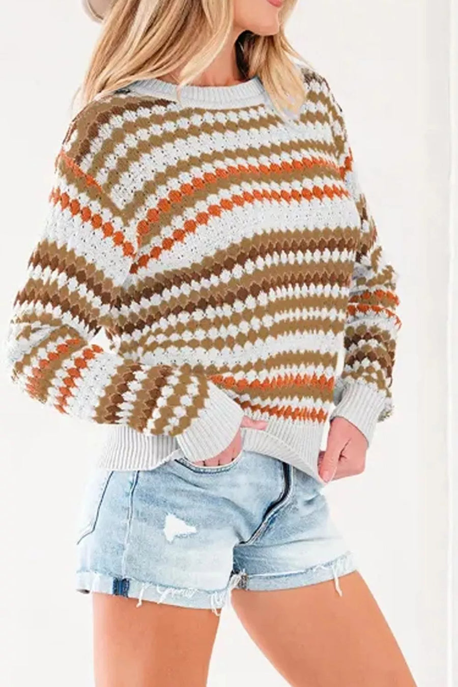 A woman wearing a chunky knit crewneck sweater with horizontal wavy stripes in white, brown, and orange, paired with light-wash denim shorts.