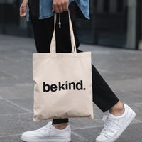 Person holding a beige tote bag with 'be kind.' text on a blurred background
