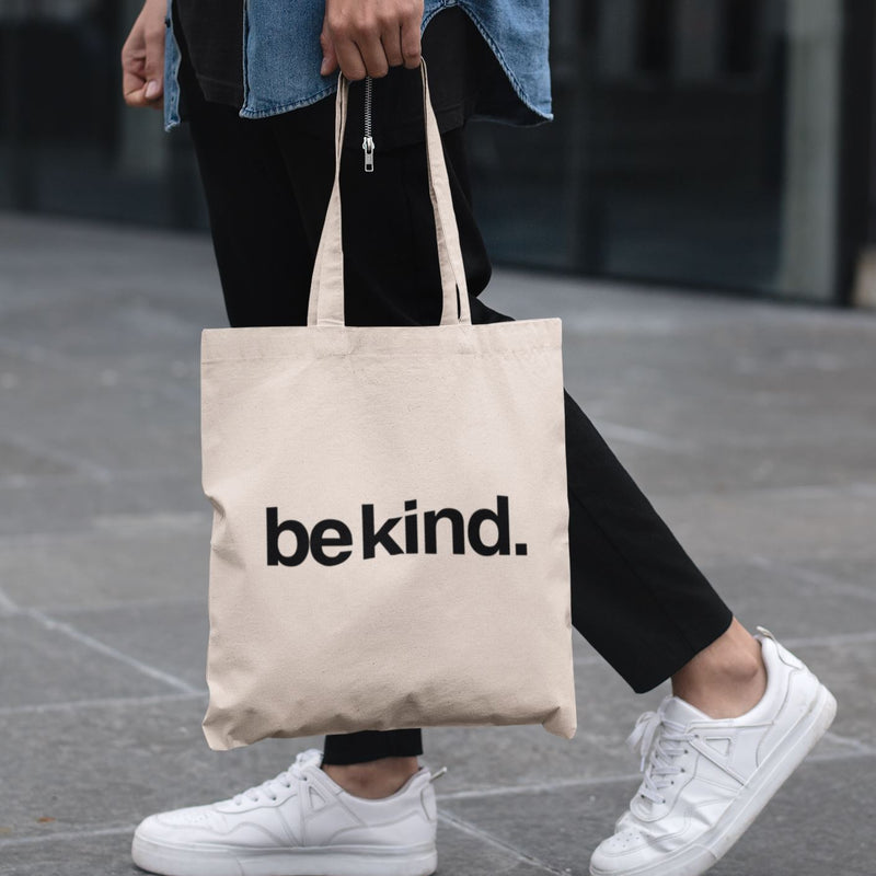 Person holding a beige tote bag with 'be kind.' text on a blurred background