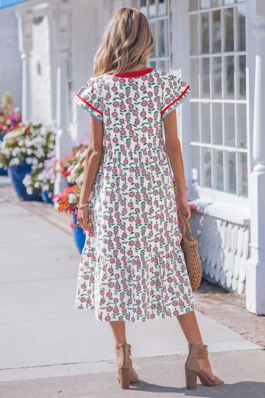 Woman wearing a floral dress standing on a sidewalk with flowers and a white building in the background.