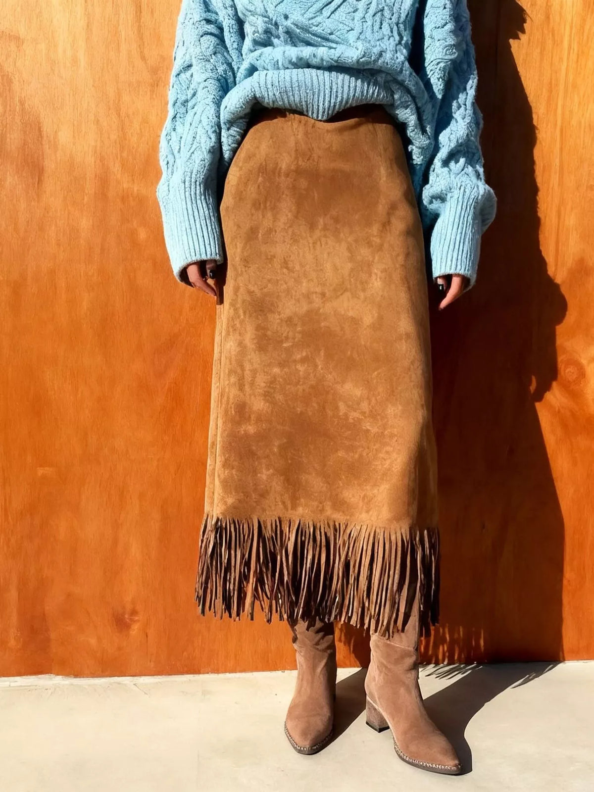 Person wearing a blue sweater and brown suede skirt with fringe against an orange wall.