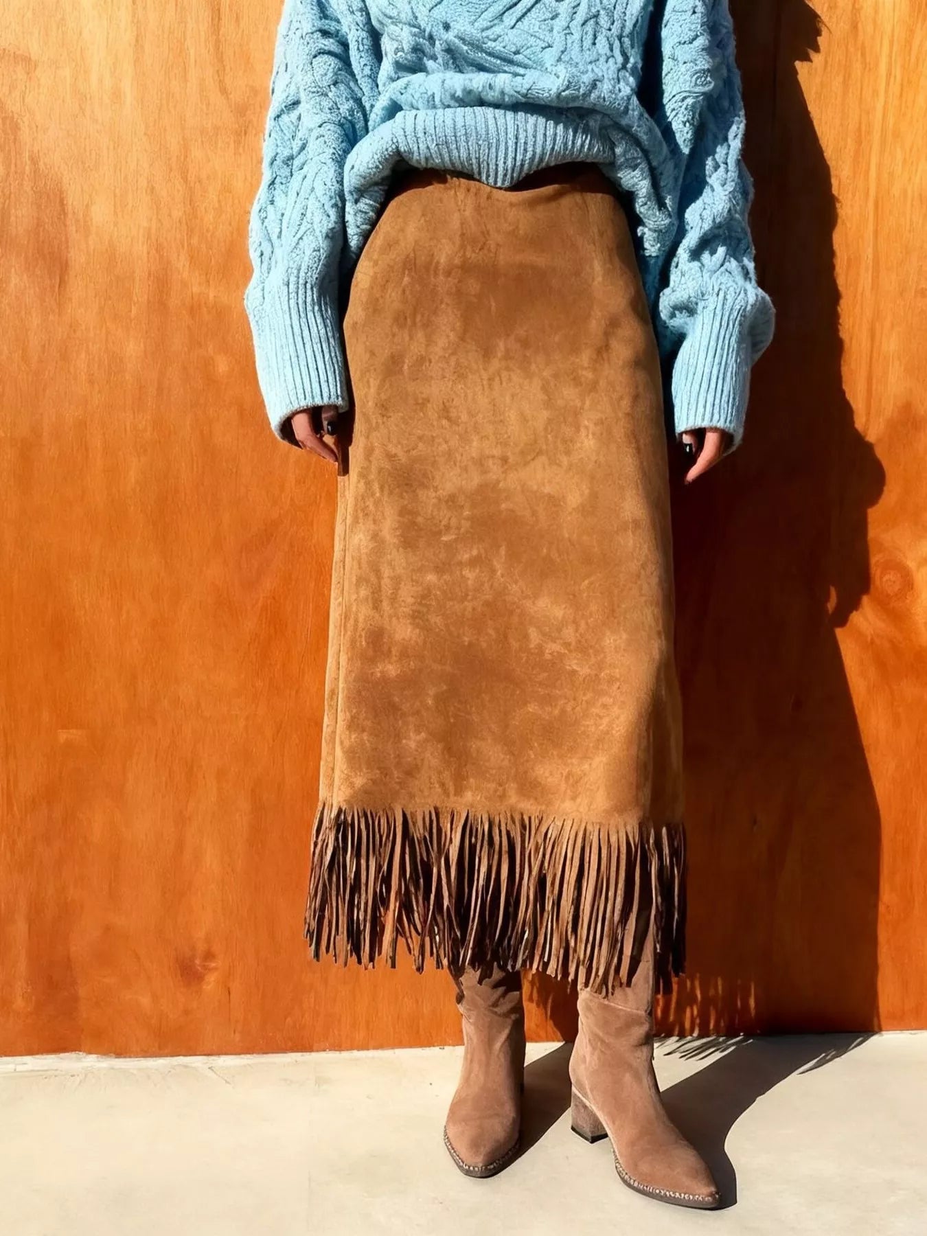 Person wearing a blue sweater and brown suede skirt with fringe against an orange wall.