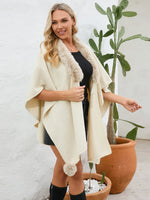 Woman wearing a cream-colored poncho with fur trim, standing in front of a white wall with plants.