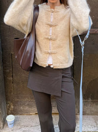 Person wearing a beige jacket with button details and dark pants, holding a brown bag.
