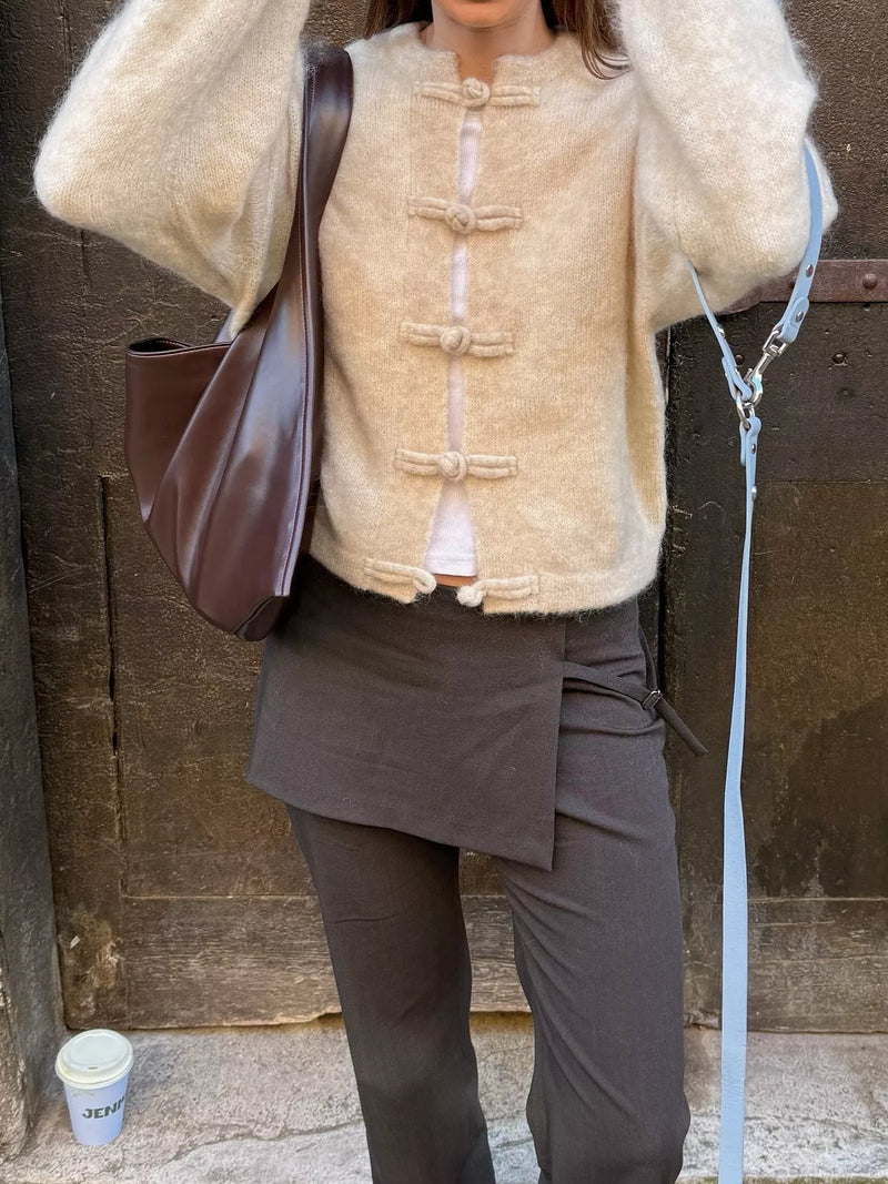 Person wearing a beige jacket with button details and dark pants, holding a brown bag.