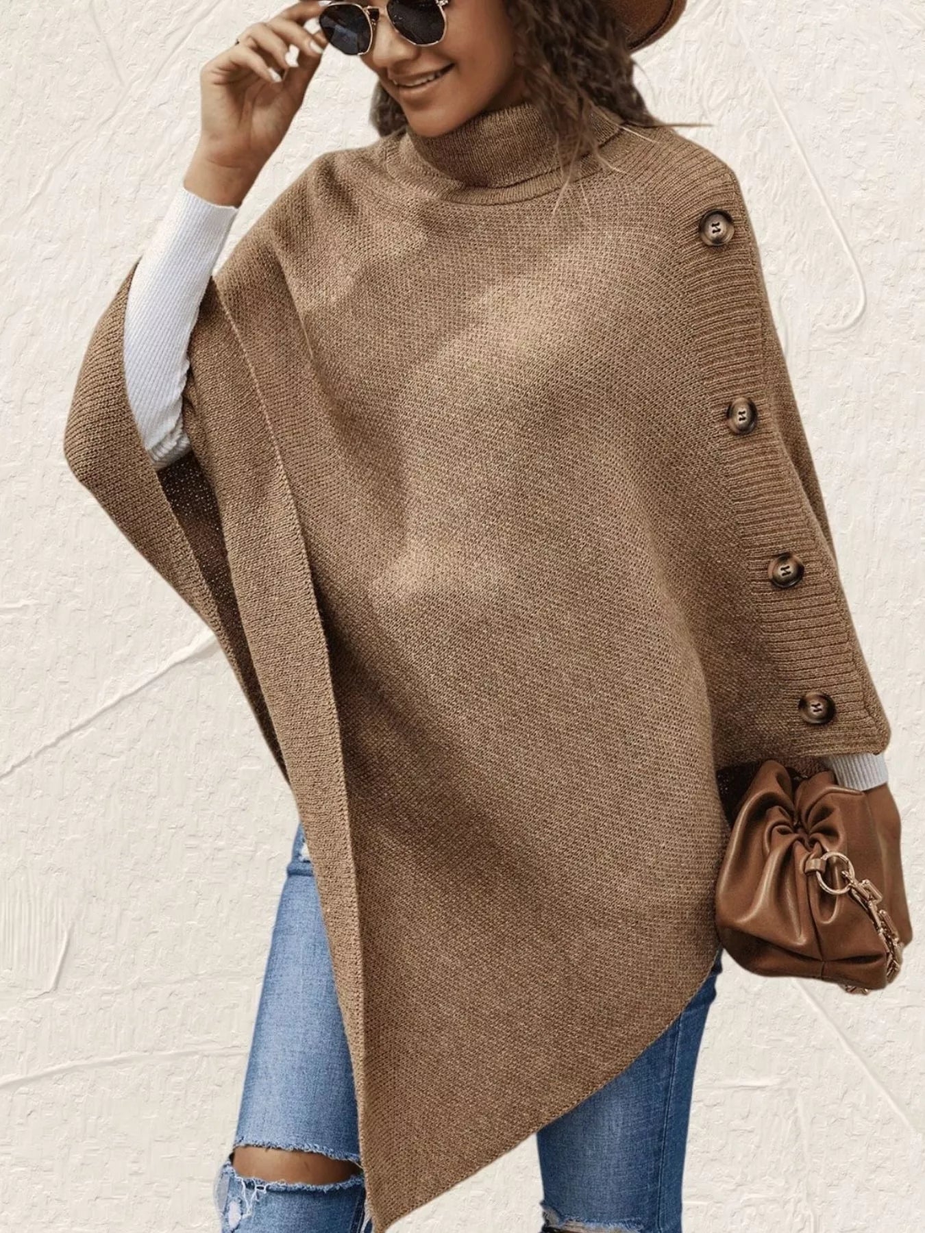 Woman wearing a brown poncho with buttons, holding a brown handbag against a light background