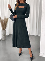 Woman wearing a dark green ribbed dress with cut-out design, holding a black handbag indoors.