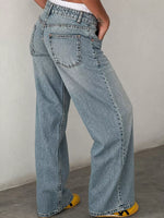 A person wearing light-wash blue wide-leg denim jeans, shown from the waist down with one hand in a pocket, standing against a plain light gray wall.