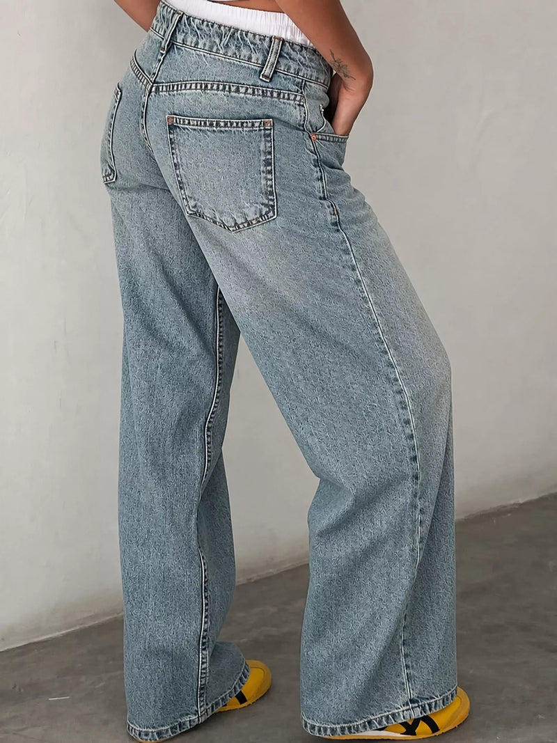A person wearing light-wash blue wide-leg denim jeans, shown from the waist down with one hand in a pocket, standing against a plain light gray wall.
