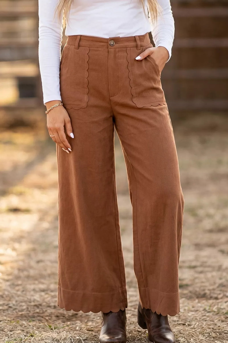 A person wearing high-waisted, wide-leg brown pants with scalloped pocket edges, styled with a white long-sleeve top and dark brown ankle boots in an outdoor setting.
