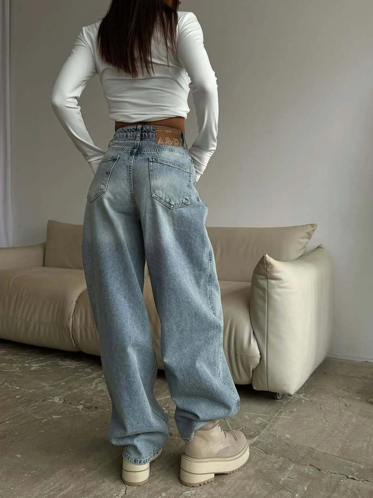 Person wearing a white long-sleeve top and blue jeans standing in a room with a beige sofa.