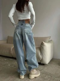 Person wearing a white long-sleeve top and blue jeans standing in a room with a beige sofa.