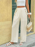 Person wearing white wide-leg pants with button details, a white top, and sandals, holding a straw bag against a beige wall.