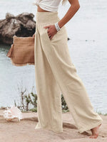 Person wearing beige wide-leg pants and a white top, holding a brown bag on a beach.
