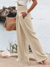 Person wearing beige wide-leg pants and a white top, holding a brown bag on a beach.