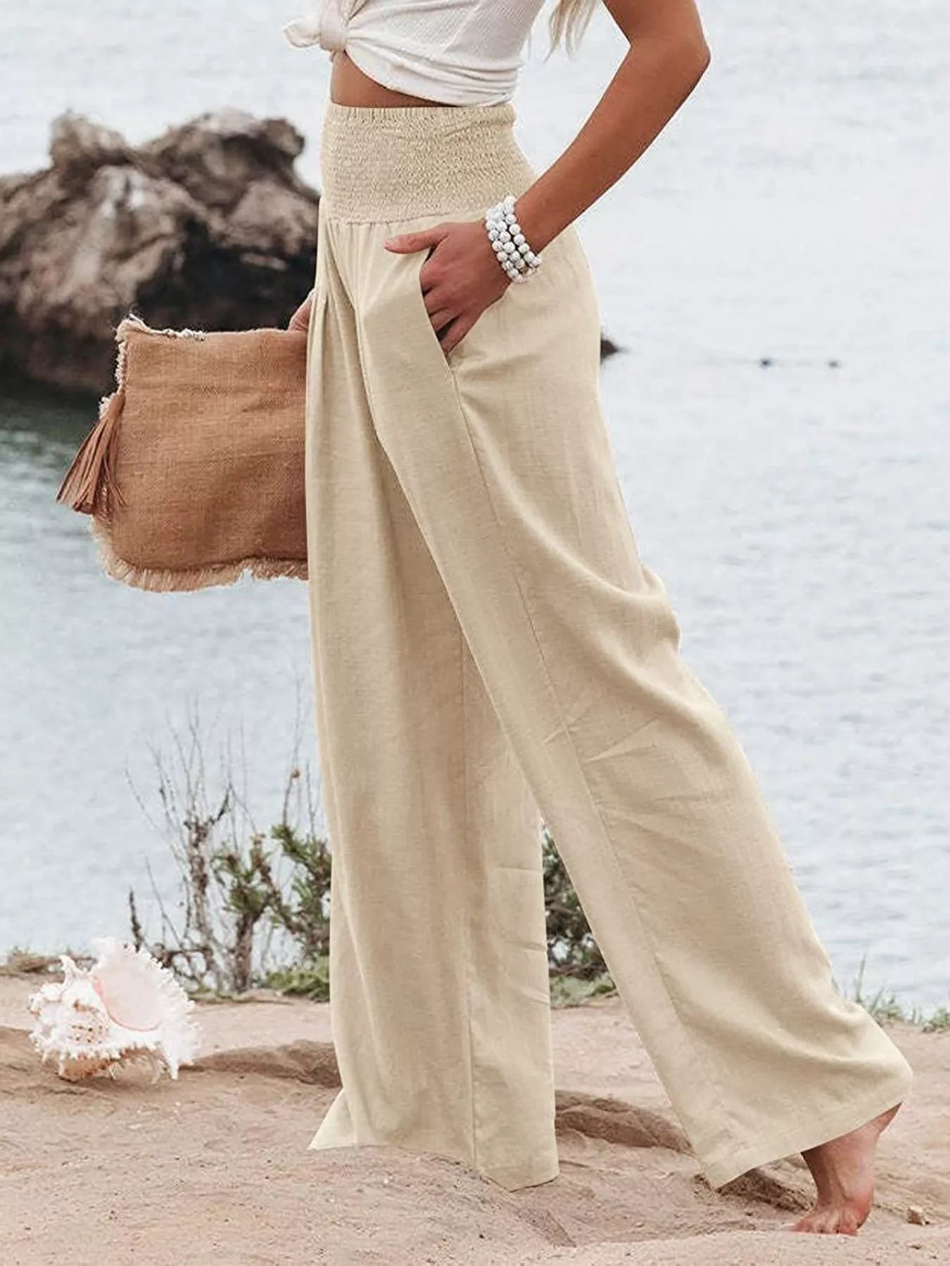 Person wearing beige wide-leg pants and a white top, holding a brown bag on a beach.