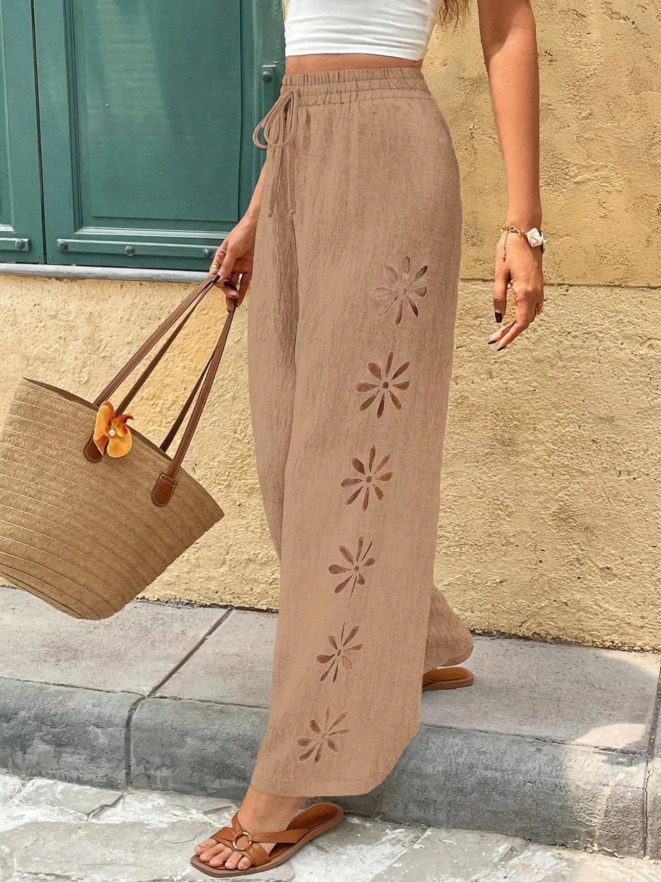 Person wearing beige pants with floral cutouts, holding a brown bag against a stone wall.