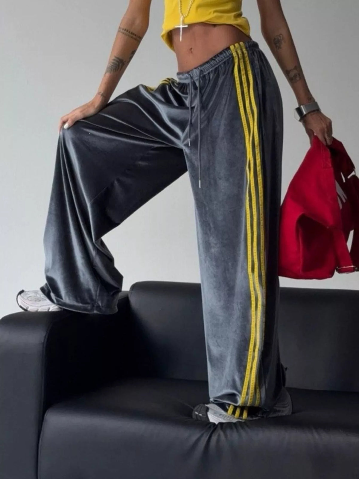 A person wearing wide-leg gray velvet sweatpants with three yellow side stripes, standing on a black leather couch and holding a red sweatshirt.