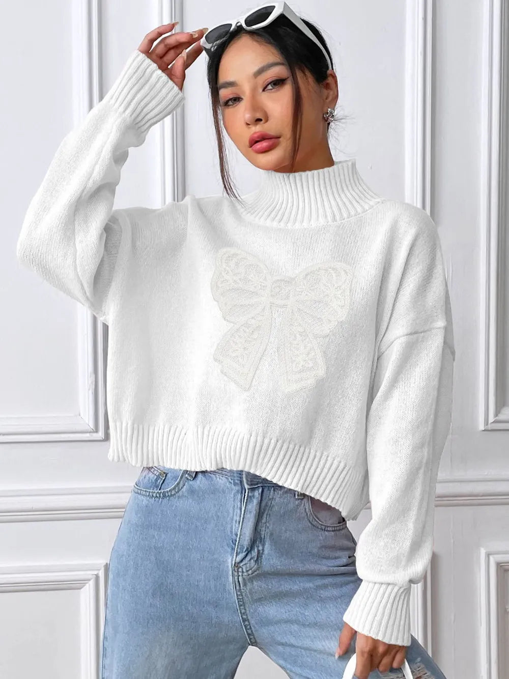 Woman wearing a white sweater and blue jeans against a white paneled wall.