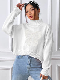 Woman wearing a white sweater and blue jeans against a white paneled wall.