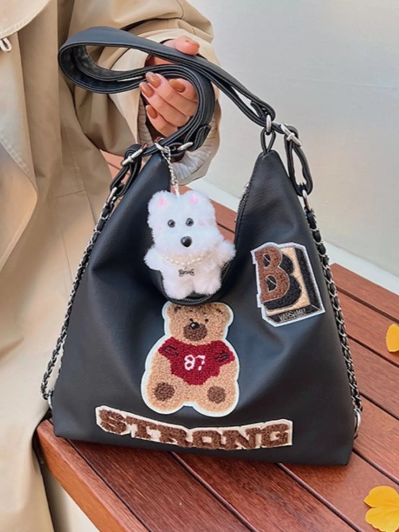 Black handbag with teddy bear and 'B Strong' patches held by a person.