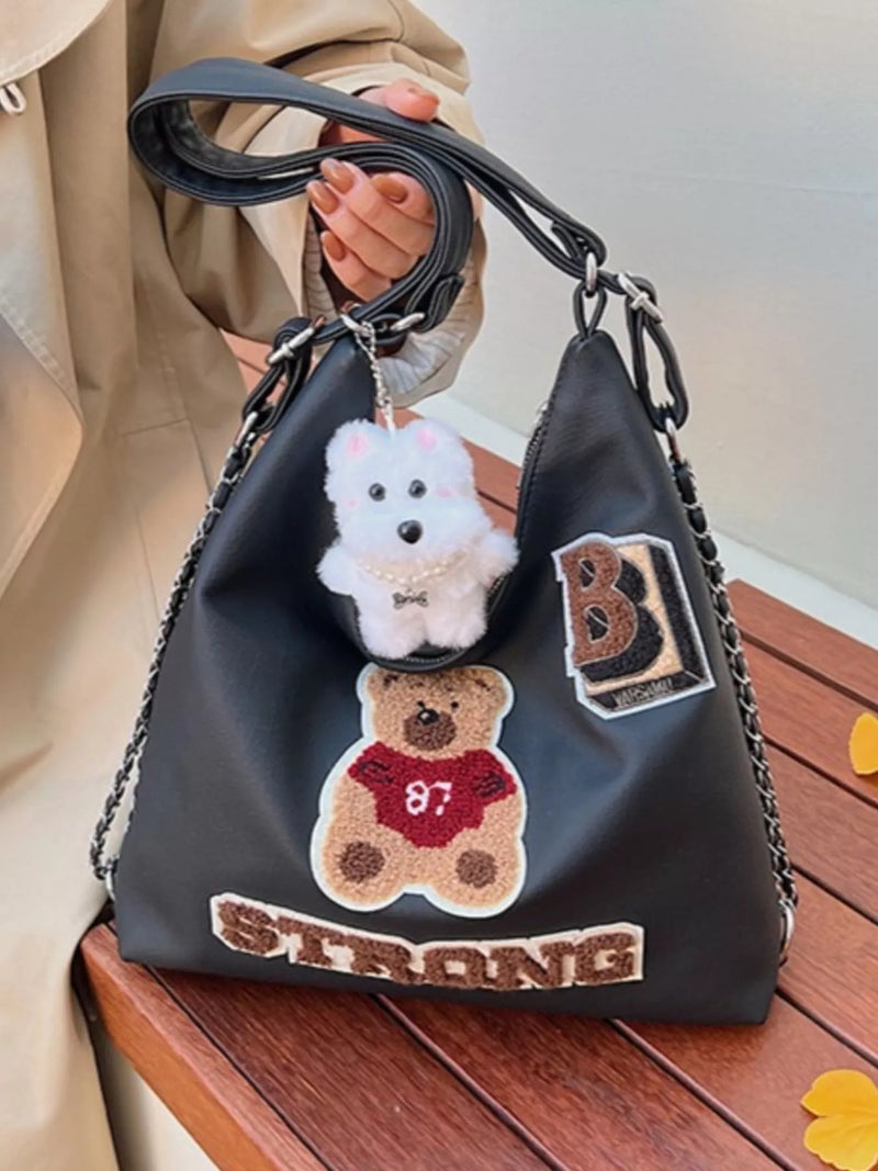 Black handbag with teddy bear and 'B Strong' patches held by a person.