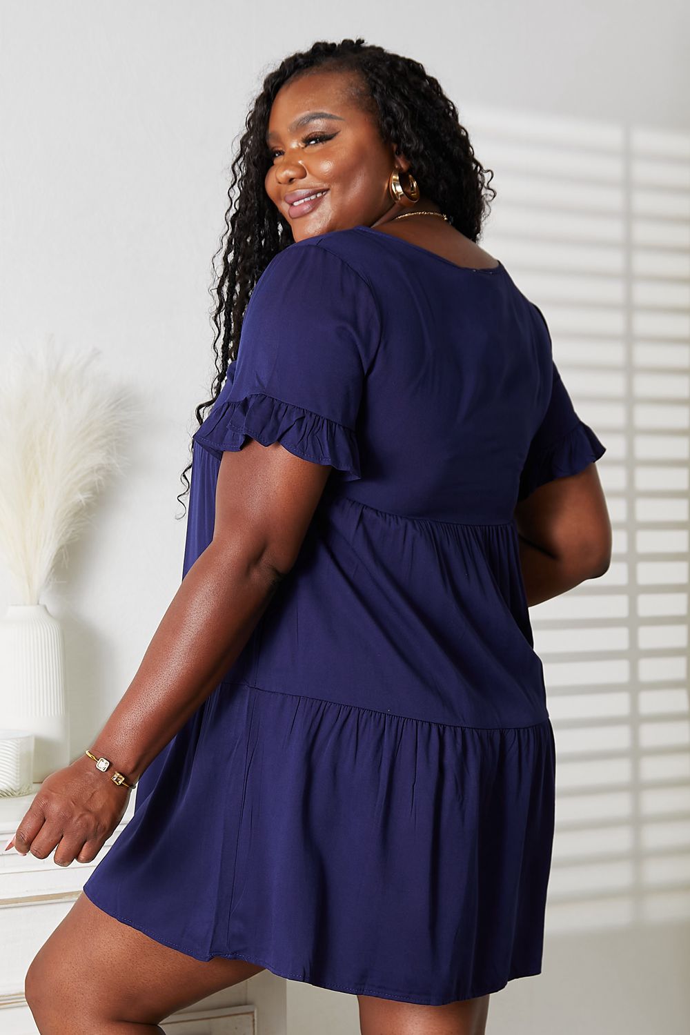 Woman wearing a blue dress with ruffled sleeves and bottom hem, standing against a neutral background.