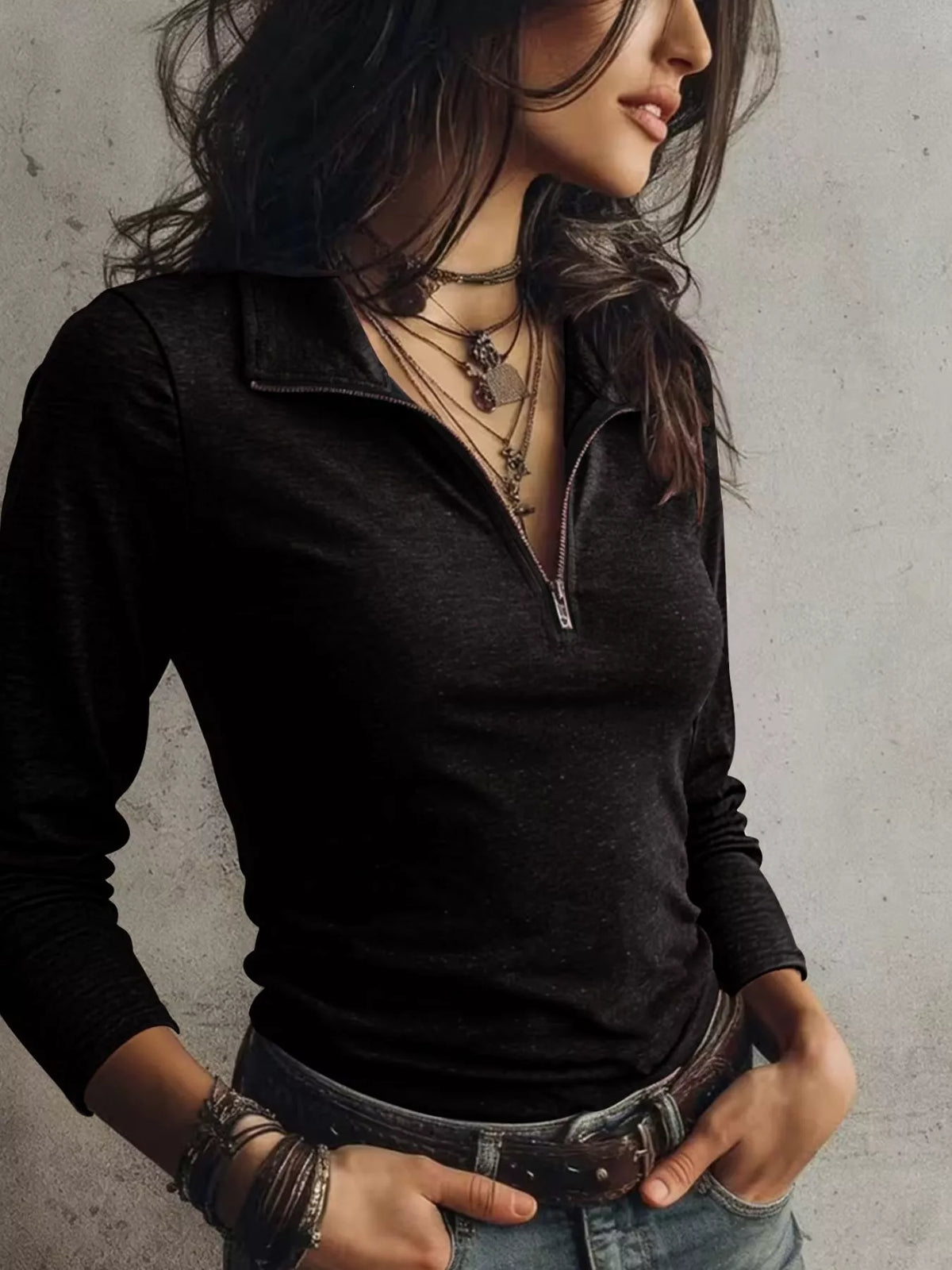 A woman wearing a black long-sleeve quarter-zip polo shirt tucked into blue jeans, accessorized with layered necklaces and bracelets, posing against a textured gray wall.