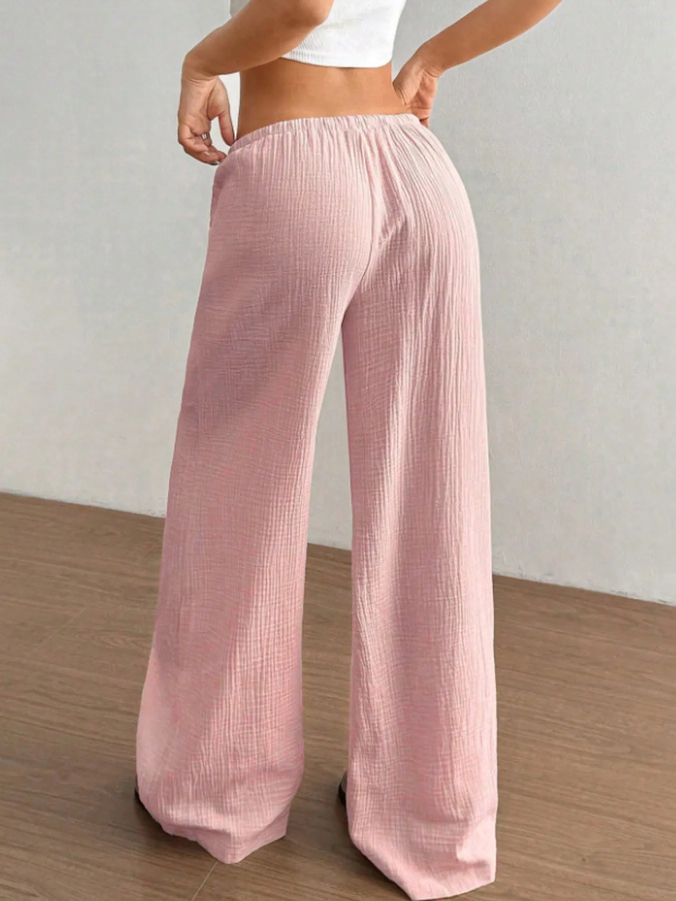 Person wearing light pink wide-leg pants on a wooden floor with a white wall background