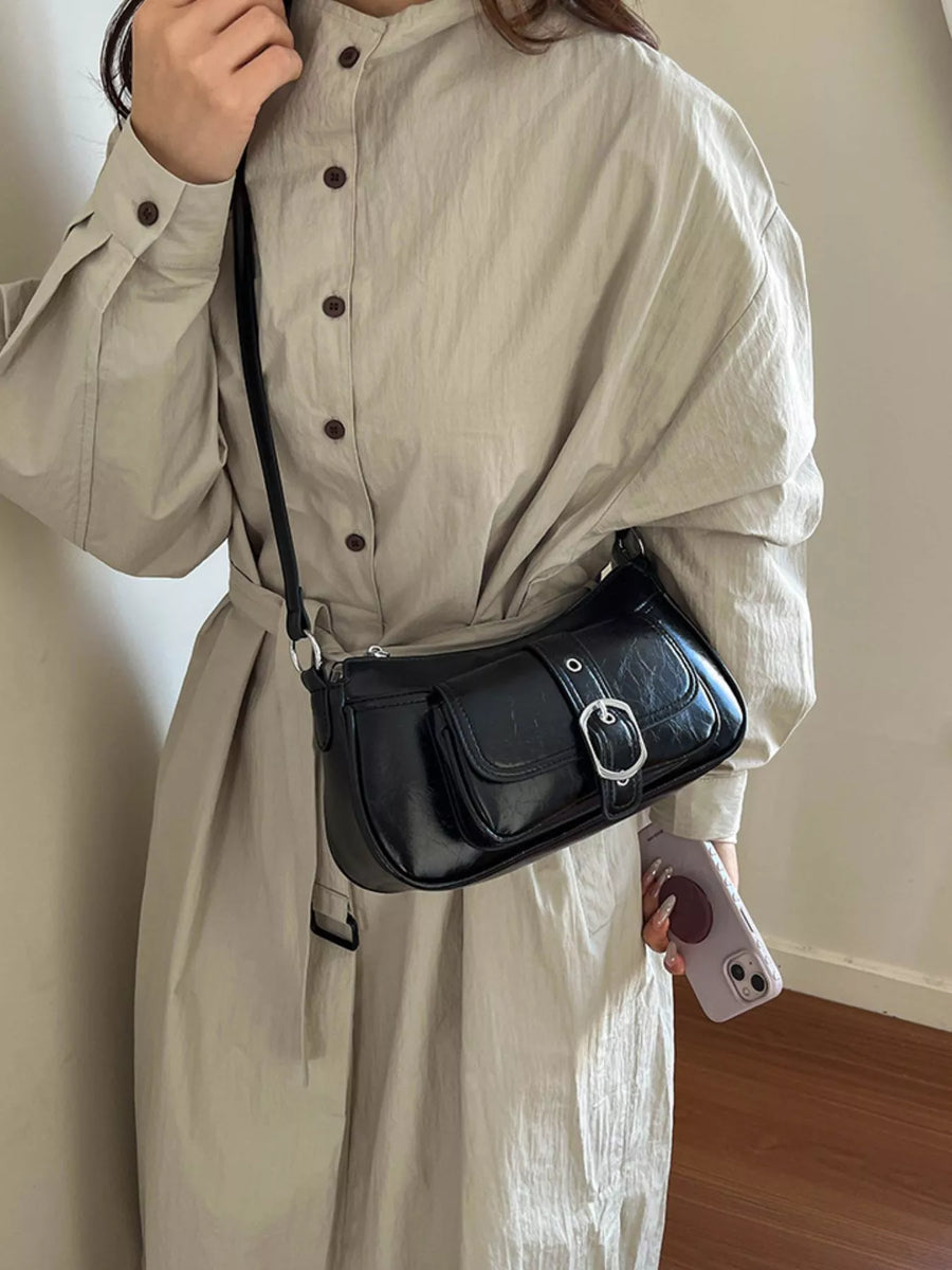 A person wearing a light beige long-sleeve button-up dress carries a black faux-leather shoulder bag with a silver buckle and strap, standing indoors against a plain wall.