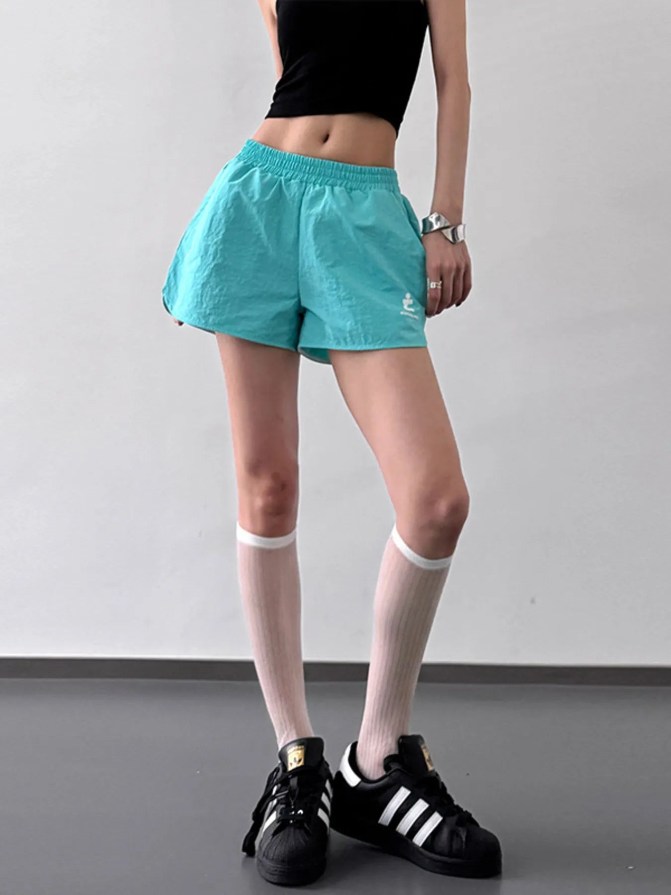 Person wearing teal shorts, black crop top, and black Adidas sneakers on a plain background