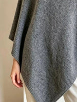A close-up of a person wearing a gray knit poncho or cape with a textured pattern, draped over the shoulders and hanging down the side, with a hand visible at the edge.