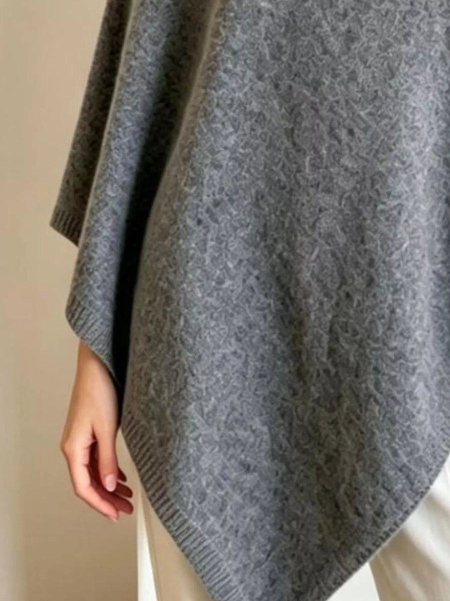A close-up of a person wearing a gray knit poncho or cape with a textured pattern, draped over the shoulders and hanging down the side, with a hand visible at the edge.