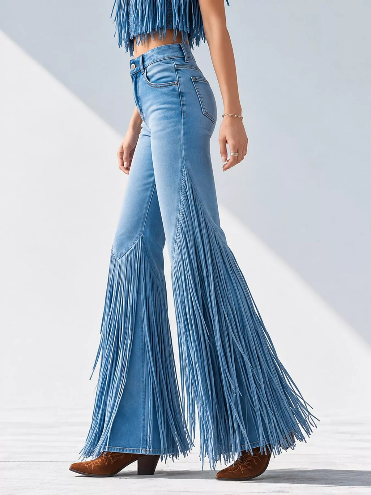 A model is shown from the waist down wearing light blue denim jeans with long fringe detailing at the hem and a matching fringe top, paired with brown leather boots, against a white background with soft diagonal shadows.