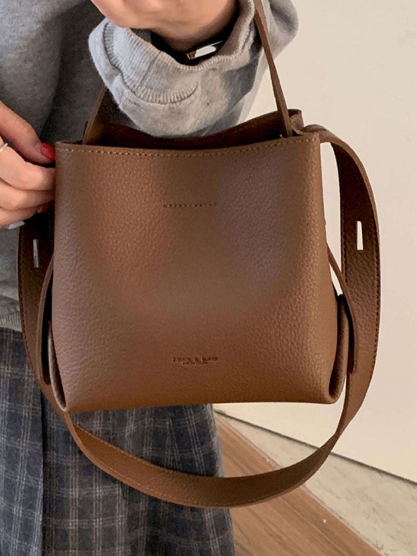 Brown leather handbag held by a person wearing a gray sweater and plaid pants.