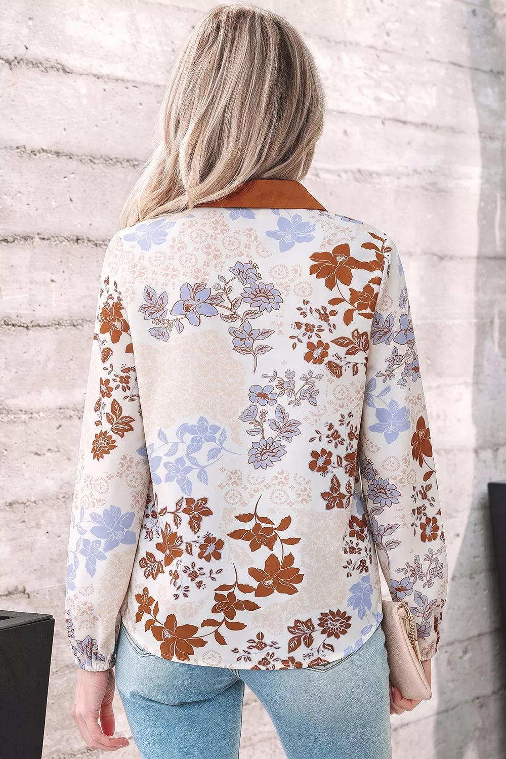 A woman viewed from behind wearing a long-sleeve blouse with a cream base and a brown, purple, and blue floral pattern, paired with light blue jeans.