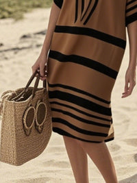 Person wearing a brown and black striped dress holding a woven bag on a beach.