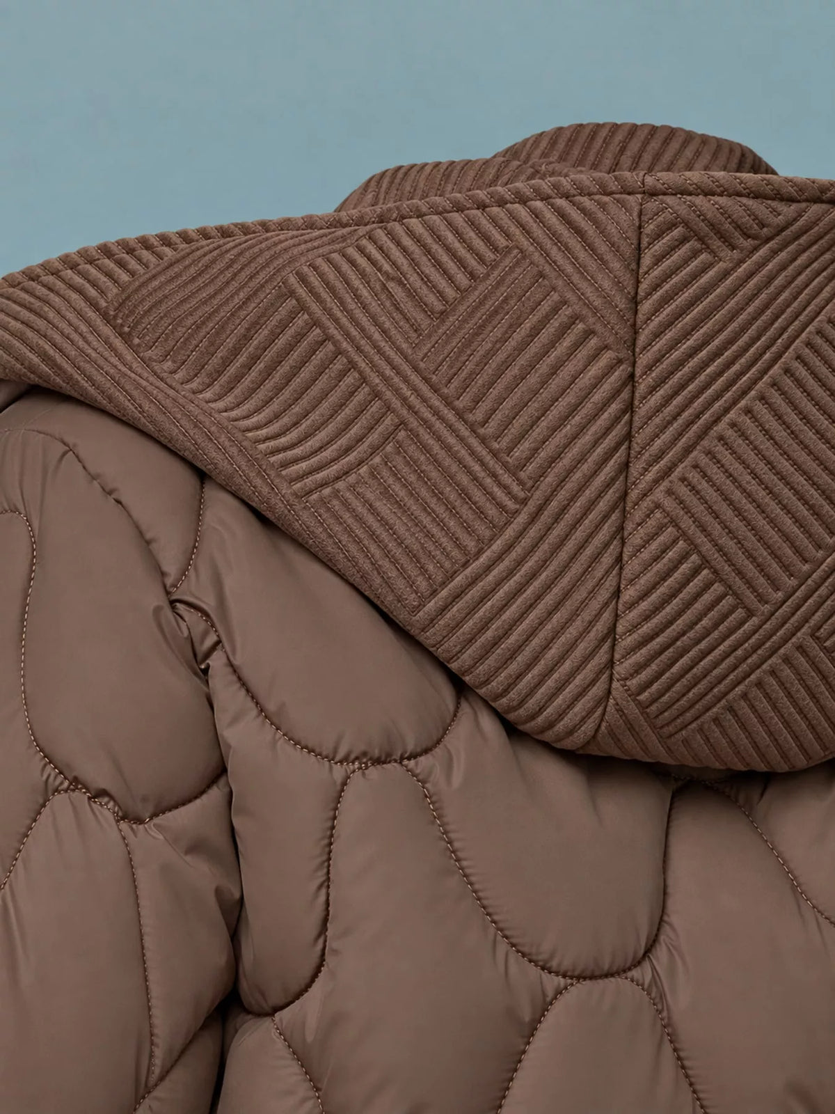 Close-up of a brown quilted puffer jacket with a ribbed hood, showing the texture and stitching details against a solid blue-gray background.