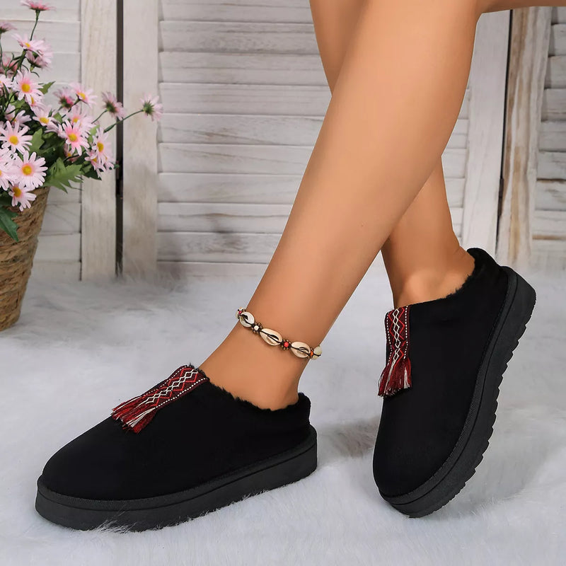 Black slip-on shoes with red and gold accents worn by a person, with a blurred background of flowers and wooden planks.