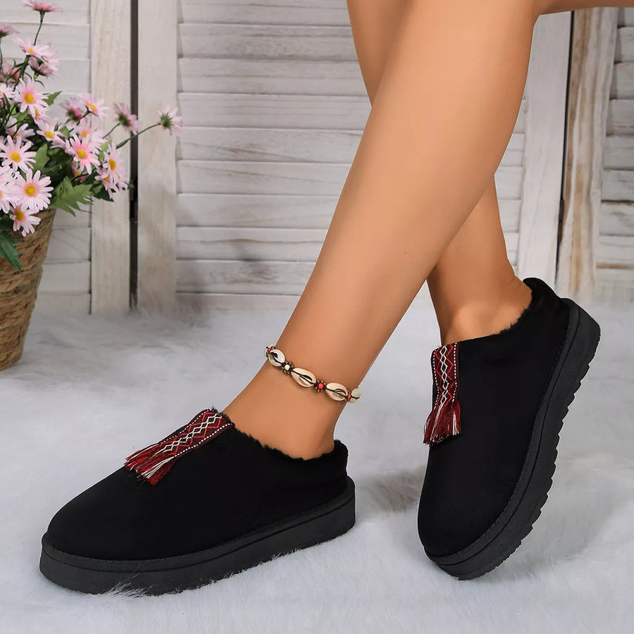 Black slip-on shoes with red and gold accents worn by a person, with a blurred background of flowers and wooden planks.