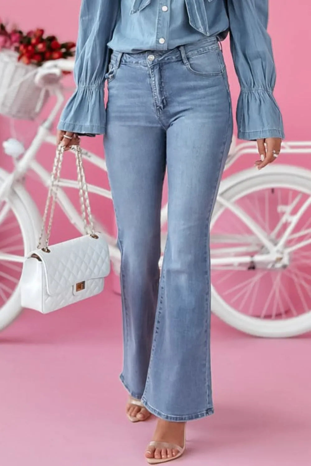 Person wearing blue jeans and a denim shirt with a white handbag, standing in front of a pink background with a bicycle.