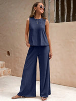 Woman wearing a blue sleeveless top and wide-leg pants standing against a beige wall.