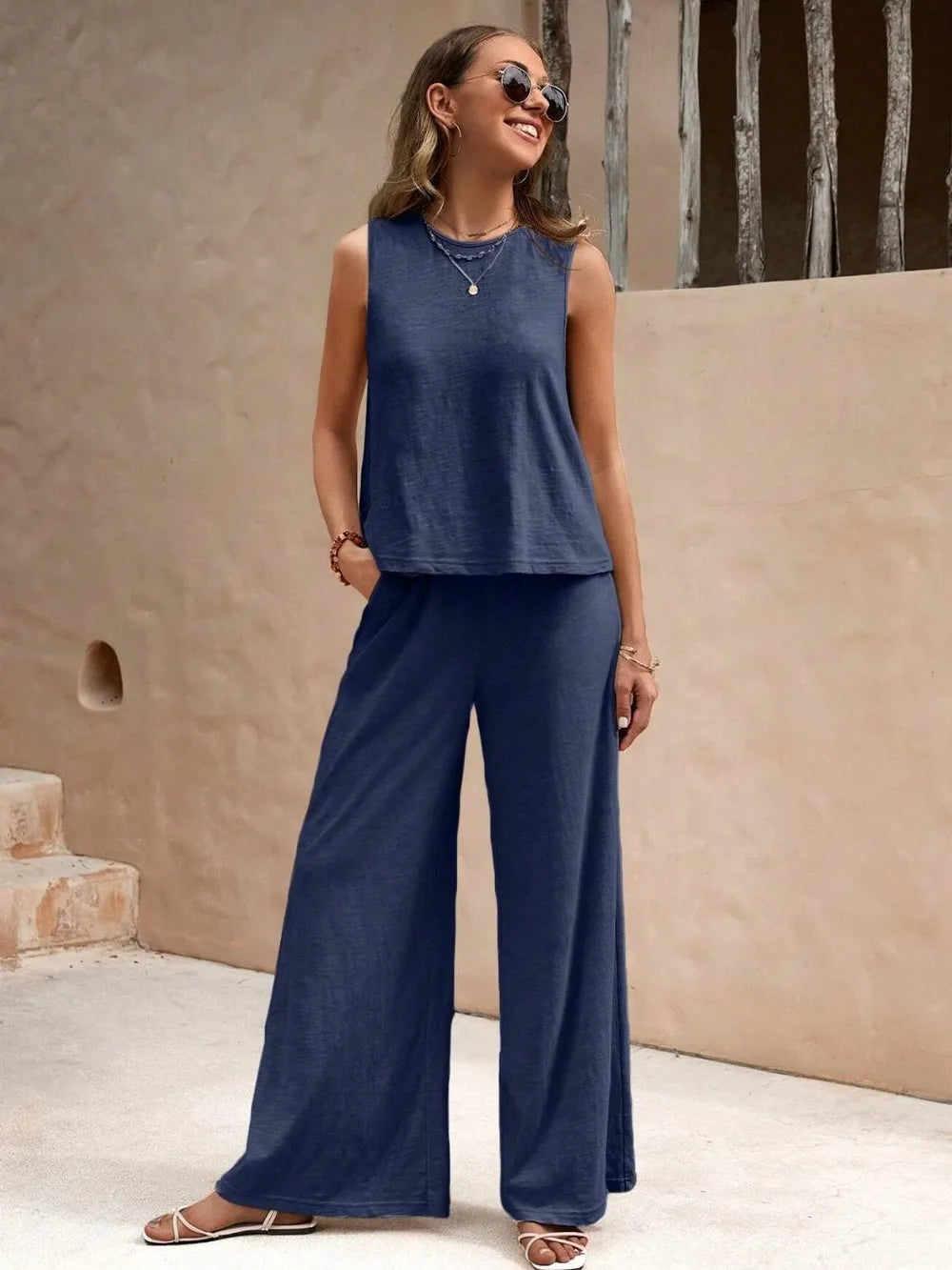 Woman wearing a blue sleeveless top and wide-leg pants standing against a beige wall.