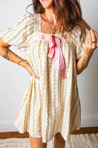 Woman wearing a floral dress with a pink ribbon, standing against a plain background.