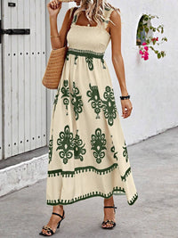 Woman wearing a beige dress with green patterns, standing against a white wall.