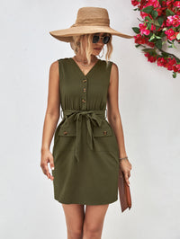 Woman wearing an olive green dress with a wide-brimmed hat and sunglasses, standing against a white background with flowers.