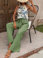 Woman sitting on a chair wearing a floral top and green pants, with a straw hat.