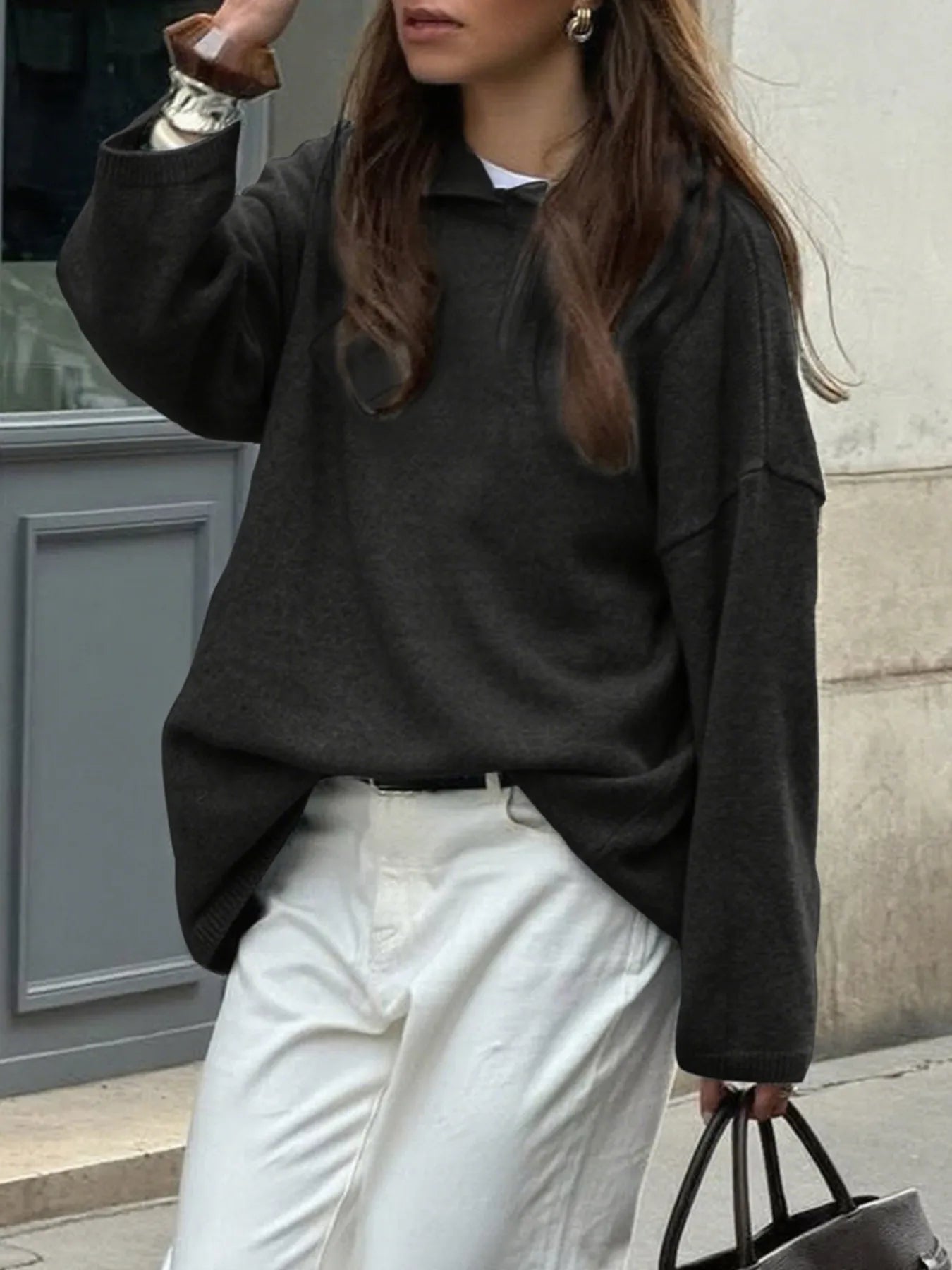 Person wearing a black sweater and white pants standing against a building wall.