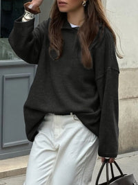 Person wearing a black sweater and white pants standing against a building wall.